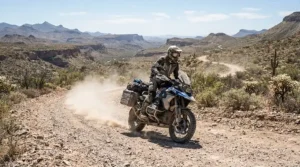 A rider wearing gauntlet gloves for adventure riding while navigating a dusty desert dirt road on a dual-sport motorcycle.